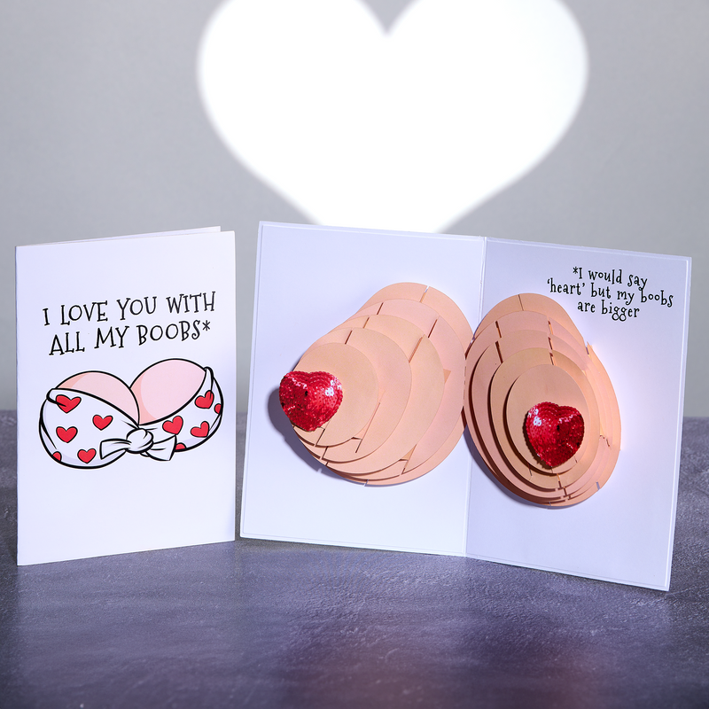 A greeting card with the text, I love you with all my boobs, and an illustration of a bra with hearts. When opened, the card pop-up of two breasts with red glittery hearts as nipples. The inside reads, I would say heart but my boobs are bigger.