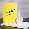 A pop-up card with a 3D hand showing the middle finger is open on a marble surface. Next to it, a bright yellow card reads Hey! in bold black letters, creating a humorous and cheeky greeting.