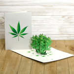 A pop-up card featuring a 3D cannabis plant is displayed open on a wooden surface, with a closed card beside it that shows a green cannabis leaf on a white background. The setting is simple and natural.