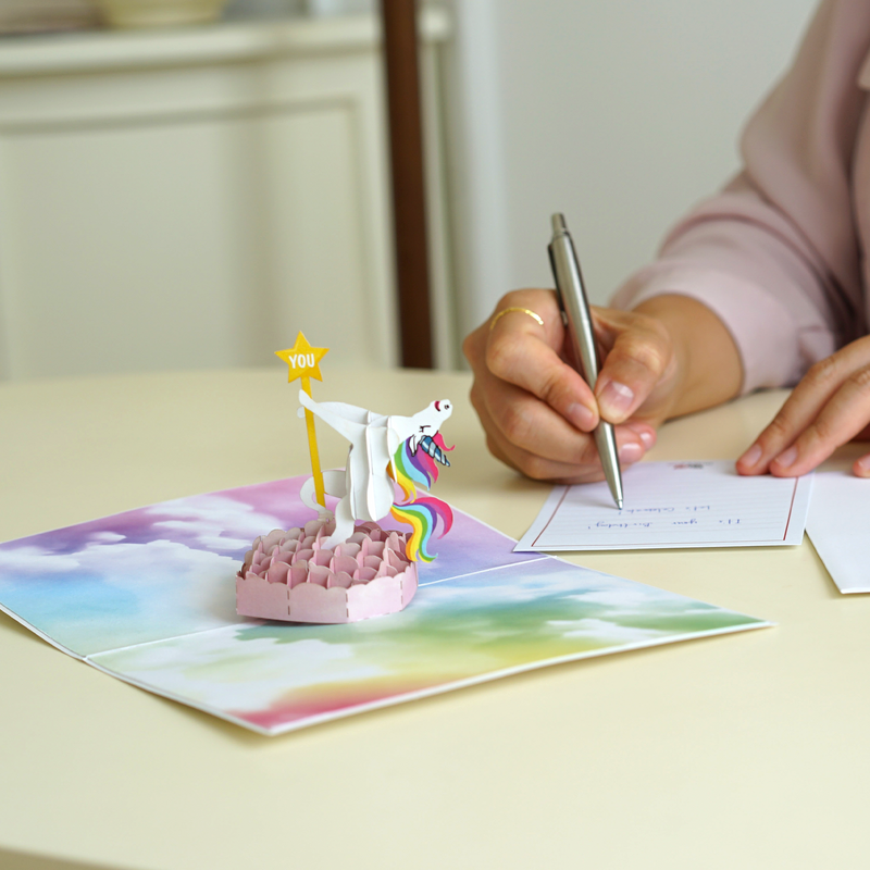 A person writes on a notecard next to an open pop-up card featuring a 3D unicorn standing on a pink platform with a rainbow mane and tail, holding a yellow star that says YOU against a colorful cloud background.