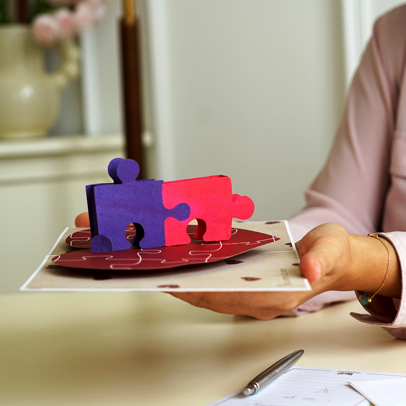 A close-up image of a person holding a pop-up card featuring two interlocking puzzle pieces, one purple and one pink, on a red base. The card is set on a desk next to a pen and a notepad, conveying a message of togetherness.