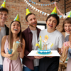 A group of five friends at a birthday party, wearing festive party hats and laughing together in a warmly lit space with brick walls and hanging bunting decorations. The woman in the center is holding an open 3D pop-up greeting card featuring a glamorous blonde woman in a white dress standing on a decorated birthday cake with a glowing striped candle. The pop-up card is vibrant with blue and gold accents, and the group appears delighted by the surprise element of the card.