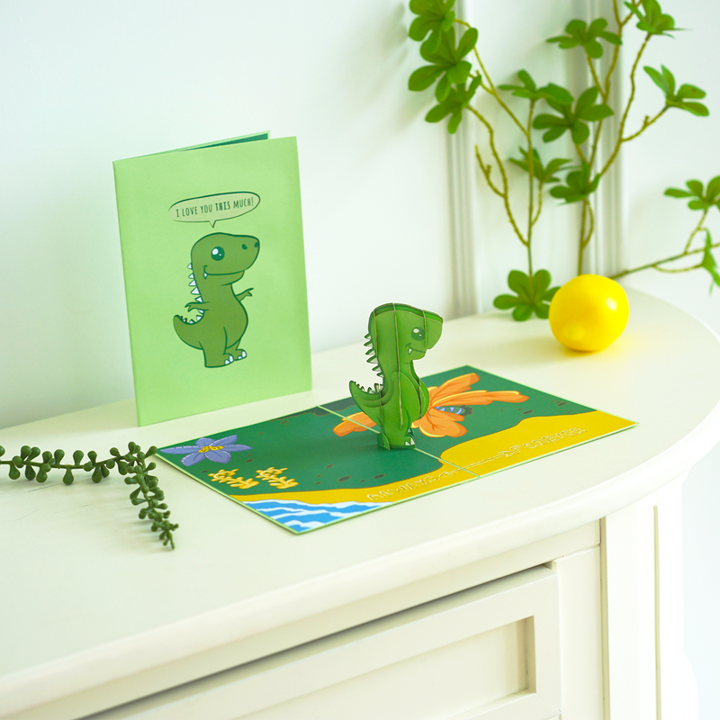 A pop-up card featuring a cute 3D green dinosaur standing on a colorful background with flowers. The matching green cover shows a dinosaur saying, I love you this much! set on a white table with plant decor, adding a playful vibe.