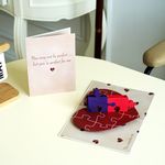 A romantic pop-up card on a table, featuring a beige cover with the text, You may not be perfect... but you're perfect for me, next to an open card with two interlocking puzzle pieces on a heart-shaped base.