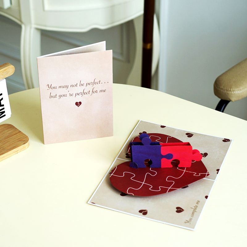 A romantic pop-up card on a table, featuring a beige cover with the text, You may not be perfect... but you're perfect for me, next to an open card with two interlocking puzzle pieces on a heart-shaped base.