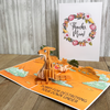 A pop-up card featuring a 3D kangaroo in an Australian landscape with the humorous text, Sorry for destroying your Down Under. In the background, a floral  Thanks, Mom! card is displayed, creating a playful contrast.