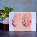 The image shows a pop-up card with a 3D illustration of breasts, featuring layered paper elements and pink nipples. The card is displayed on a gray surface with a potted plant in a pink ceramic pot beside it.