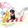 A pop-up birthday card featuring three playful dogs wearing party hats, surrounded by balloons and musical notes. The card includes the message, "Nope. I haven’t SEEN any CAKE!" adding a humorous touch to the festive design.