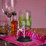 A pop-up card featuring a dancer on a pole is placed on a sparkly surface with scattered confetti. In the background, there are glasses, a mini bottle of liquor, and a striped pink backdrop, creating a celebratory scene.