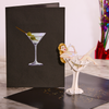 A pop-up card featuring a 3D woman lounging in a martini glass next to a black cover card with a martini glass and two olives. The setting is on a marble surface with colorful decorations in the background, adding a festive touch.