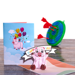 A pop-up card featuring a smiling piggy bank holding a HAPPY BIRTHDAY! banner is in the foreground. A closed card with a flying pig holding balloons is next to it. In the background, a dartboard with red darts is visible.