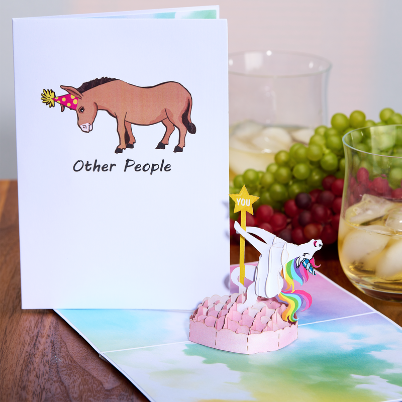 A pop-up card shows a unicorn with a rainbow mane holding a YOU star, standing on a pink platform. Next to it, a closed card displays a donkey wearing a party hat with the text  Other People.  Drinks and grapes are in the background.