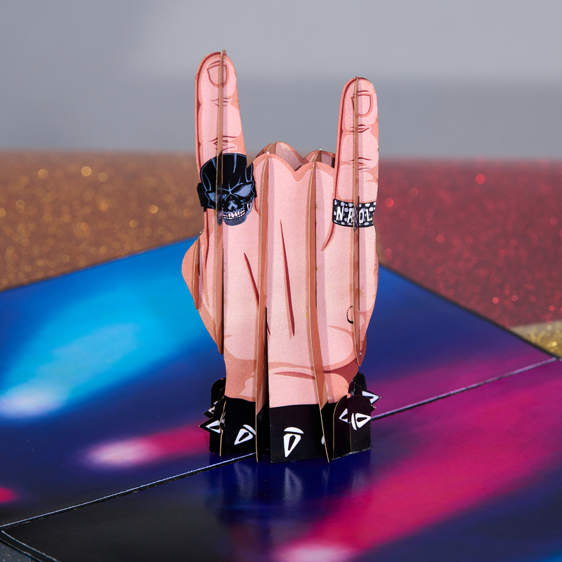 A pop-up card featuring a 3D hand making the rock 'n' roll gesture, with skull and studded rings, set against a vibrant, colorful stage backdrop with glittery details, creating a dynamic and festive vibe.