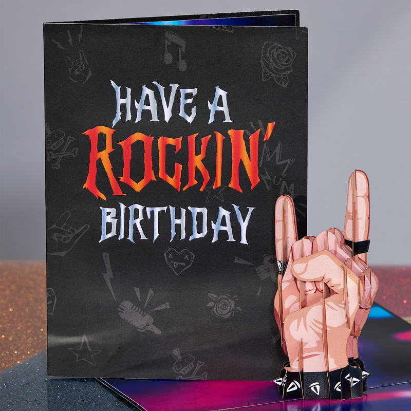 A close-up of a pop-up card showing a 3D hand making the rock 'n' roll sign, complete with wristbands and studs, set against a colorful stage backdrop. Part of the Have a Rockin' Birthday card cover is visible in the background.