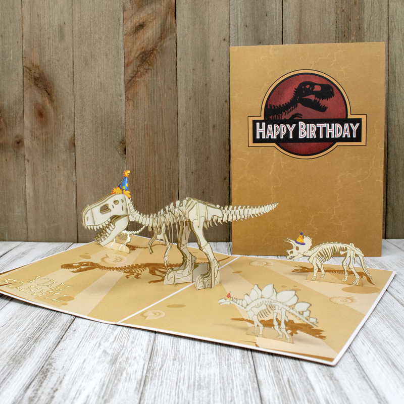 A pop-up card featuring dinosaur skeletons, including a T-Rex and Triceratops, both wearing party hats, is displayed open against a wooden background. The card cover says Happy Birthday with a T-Rex silhouette.