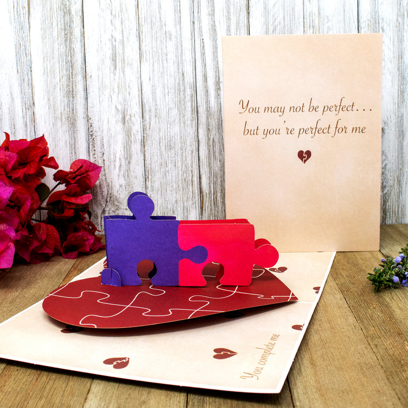 A pop-up card with interlocking blue and red puzzle pieces on a heart-shaped base, set against a wood background. The card's cover reads, You may not be perfect... but you're perfect for me, with red flowers in the background.