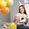 A smiling girl in a party hat holds a "We heard there was cake!" birthday card, with balloons and a wrapped gift nearby. A sleepy puppy rests on the couch, adding to the cozy celebration.