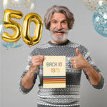 Smiling man in a festive sweater holds a "Back in 1975" card, with gold "50" balloons and confetti balloons in the background.