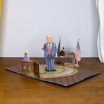 A pop-up greeting card featuring a cartoon figure resembling Donald Trump giving a thumbs-up while standing in a depiction of the Oval Office. The background includes a desk, flags, and a Happy Birthday cake.