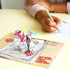 A person writes on a notecard next to a pop-up card featuring cartoon plugs with cheerful expressions and speech bubbles saying Have a super-charged day! and Keep Going!  The card says, Nothing but good vibes coming your way.