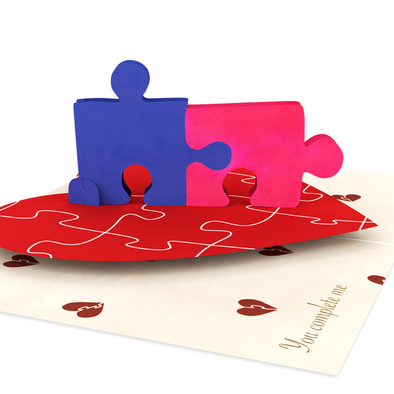 A pop-up card featuring two interlocking puzzle pieces, one blue and one pink, on a red heart-shaped base. The background has small heart illustrations and the text You complete me is written in the corner.