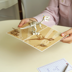 A person holds an open pop-up card featuring a T-Rex skeleton wearing a party hat, with fossil illustrations on a beige background. The card is positioned above a table with a handwritten note and a pen nearby.