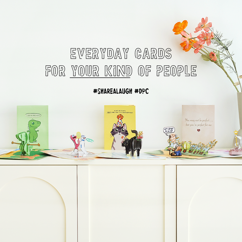 A collection of colorful and humorous pop-up greeting cards displayed on a white cabinet, with the slogan 'Everyday Cards for Your Kind of People' above them. Hashtags #ShareALaugh and #DPC are also featured, along with a vase of flowers