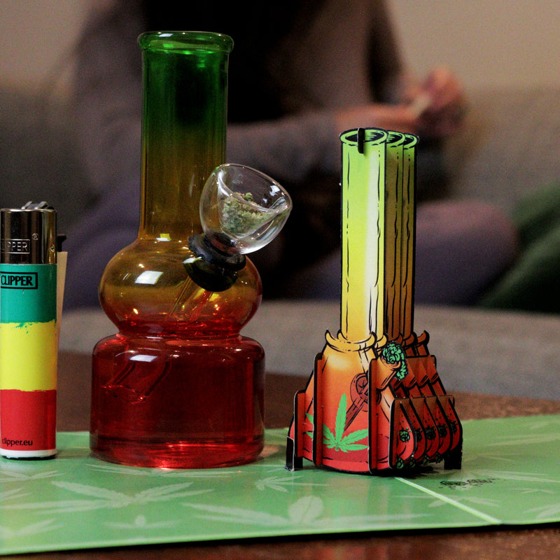 A pop-up card featuring a 3D bong design stands next to a real glass bong filled with herbs and a lighter in Rasta colors. The background shows a blurred person, adding a casual, relaxed atmosphere to the scene.