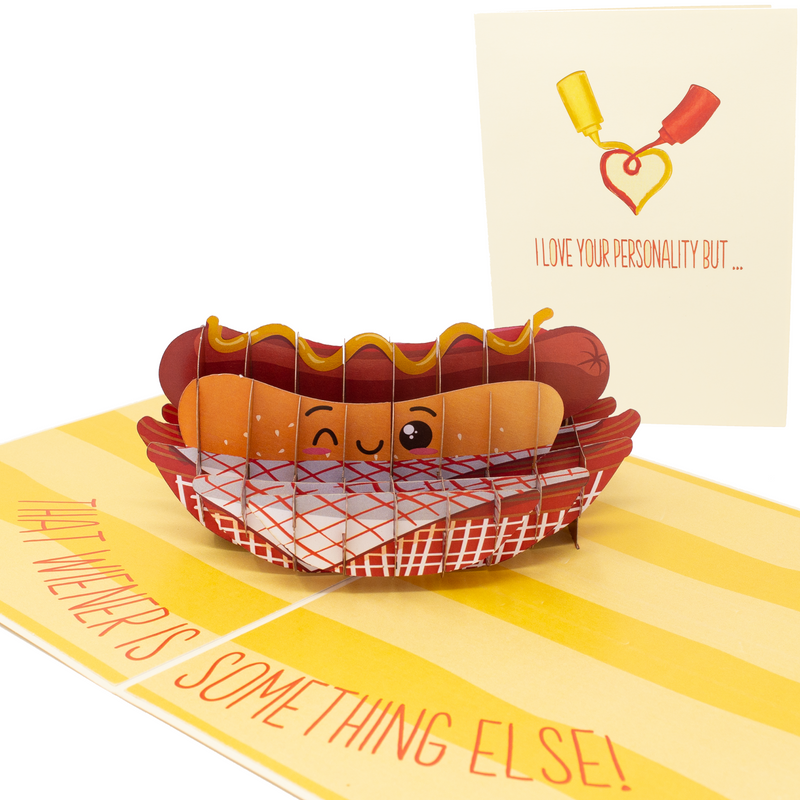 The image shows a humorous pop-up card featuring a smiling hot dog in a basket. The card's cover reads, I love your personality but... with mustard and ketchup forming a heart, and inside, it says, That wiener is something else!