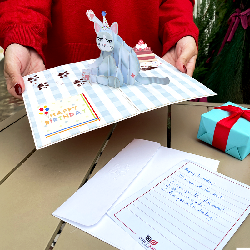 A 3D pop-up birthday card featuring a grumpy cat in a party hat on a checkered background. A handwritten note and white envelope sit nearby, with a blue gift box wrapped in a red ribbon. A person in a red sweater holds the card in a festive setting with greenery.