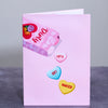 A pink greeting card cover featuring a box of candy hearts with playful phrases. Three hearts spill out, reading Let's Get Naked, adding a cheeky and romantic twist to the design.