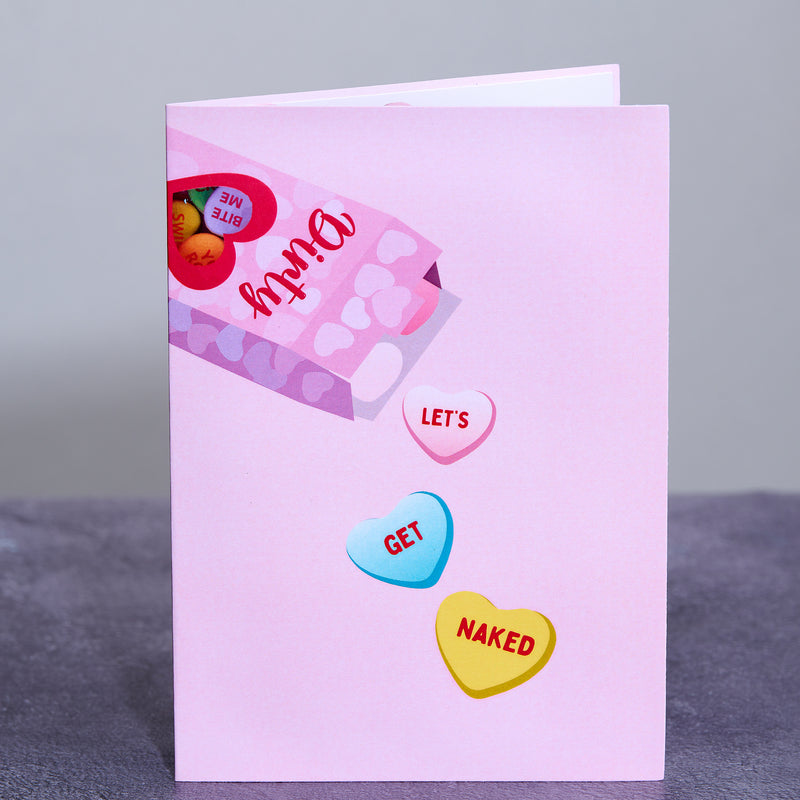 A pink greeting card cover featuring a box of candy hearts with playful phrases. Three hearts spill out, reading Let's Get Naked, adding a cheeky and romantic twist to the design.