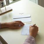 A person writes a message on a notepaper that reads Happy Birthday, You Are Amazing, with an envelope nearby on the table. The envelope is labeled as 7 inches wide and 5 inches tall, and the notepaper is from DirtyPop, adding a personalized touch