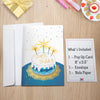 A flat-lay display of a birthday pop-up card set on a wooden surface, accompanied by an envelope and an informational card listing the contents: '1 – Pop Up Card 8” x 5.5”, 1 – Envelope, 1 – Note Paper.' The card cover features an illustration of a festive birthday cake with golden sparklers, a blue and gold base, and the words 'Happy Birthday' written in gold script. Surrounding the setup are party elements, including a gold-wrapped gift, pink striped candles, confetti, and a plate with a cupcake and small