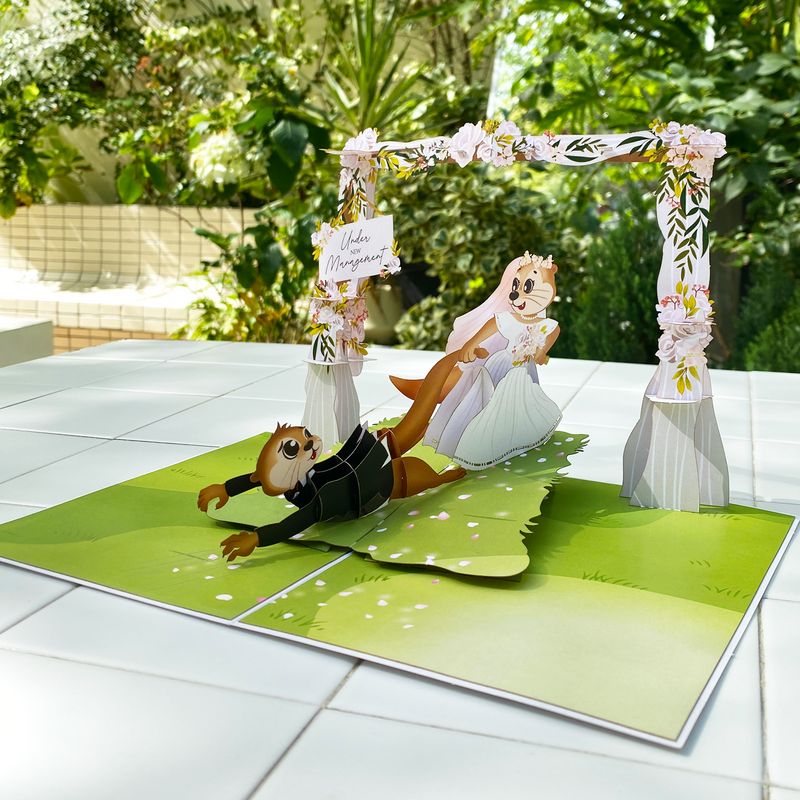 A 3D pop-up wedding card displayed on a white tiled surface with lush greenery in the background. The card features two cartoon otters in wedding attire, with the bride wearing a white gown and flower crown, playfully pulling the groom by his leg as he falls backward in a tuxedo. The scene is set under a floral arch adorned with pink and white flowers, with a sign that reads 'Under New Management.' The card's base is designed to resemble a grassy outdoor setting with scattered pink petals