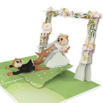 A 3D pop-up greeting card featuring two cartoon otters in wedding attire under a floral arch. The bride otter, wearing a white dress and a flower crown, is playfully pulling the groom otter, dressed in a tuxedo, by his leg as he falls backward. The arch is decorated with pastel flowers and greenery, with a small wooden sign that reads 'Under New Management.' The scene is set on a grassy surface with scattered pink flower petals