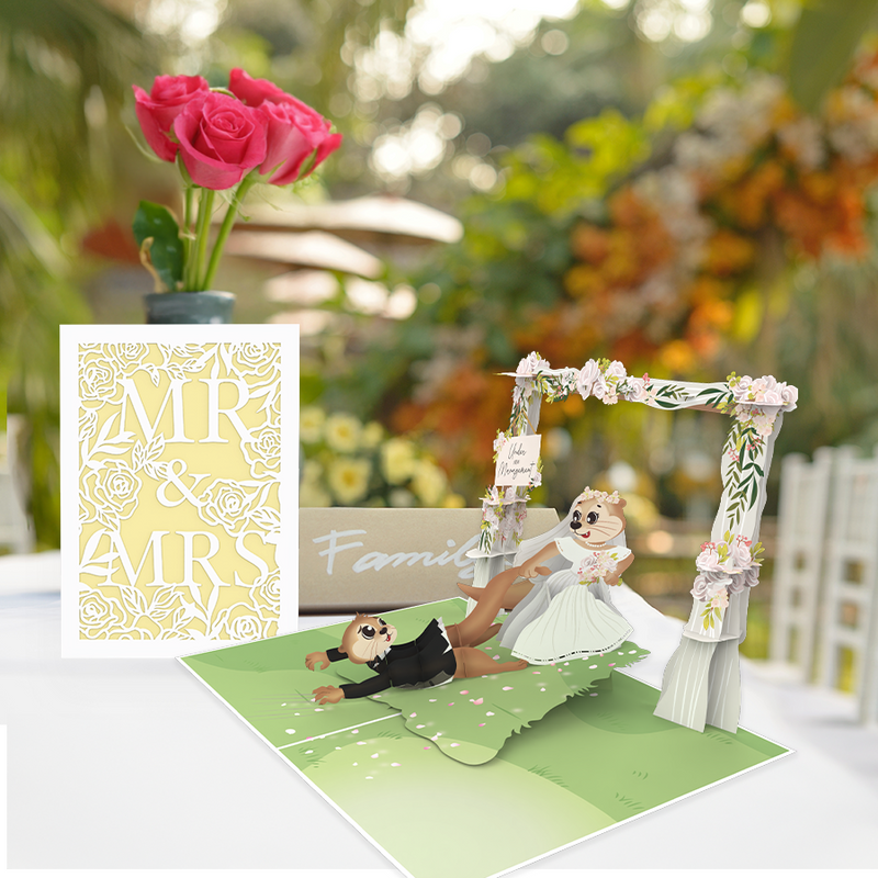 A wedding-themed pop-up greeting card displayed on a table with a blurred outdoor wedding setting in the background. The card features two cartoon otters in wedding attire, with the bride playfully pulling the groom by his leg under a floral arch that has a sign reading 'Under New Management.' Next to it is a flat card with an intricate laser-cut design that reads 'MR & MRS' on a soft yellow background. A vase with pink roses adds a romantic touch to the scene.