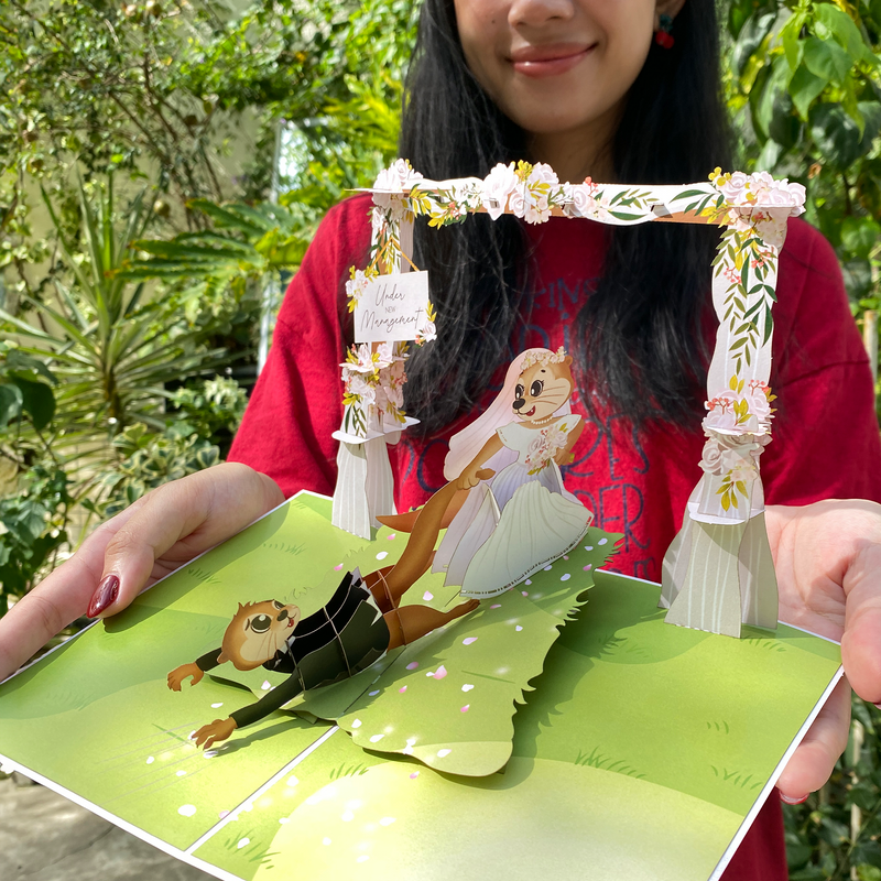 A person wearing a red shirt holds an open 3D pop-up greeting card featuring two cartoon otters in wedding attire. The bride otter, dressed in a white gown and flower crown, playfully pulls the groom otter, who is wearing a tuxedo, by his leg as he falls backward. The scene is set under a floral wedding arch with a small sign that reads 'Under New Management.' The background includes lush greenery, suggesting an outdoor setting.