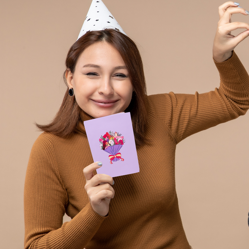 A woman wearing a brown sweater and a white party hat with black stars smiles while holding a purple greeting card with a colorful illustration of a bouquet tied with a red and yellow ribbon.