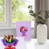 A purple greeting card with a colorful bouquet illustration is displayed on a table near a white vase with green leaves, with an open pop-up card version of the bouquet in front of it.