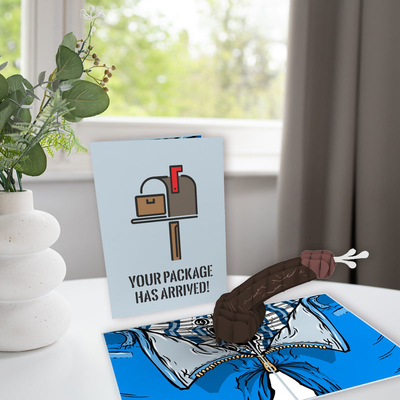 A 3D pop-up card on a table with a humorous object resembling a sausage emerging from a blue background. Behind it, a light blue greeting card reads YOUR PACKAGE HAS ARRIVED! next to a white vase with greenery.