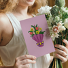 A woman in a white dress holds a bouquet of fresh flowers in one hand and a pink greeting card with an illustration of a colorful bouquet wrapped in purple paper with a yellow and purple ribbon in the other.