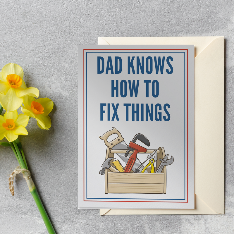 A greeting card with the text DAD KNOWS HOW TO FIX THINGS in bold blue letters, featuring a toolbox with tools, placed on a gray surface next to a yellow flower and a matching envelope.