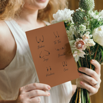 A woman in a white dress holds a bouquet of flowers and a brown greeting card featuring simple line drawings of different breast shapes labeled with humorous terms like Boobies and Ta-Tas.