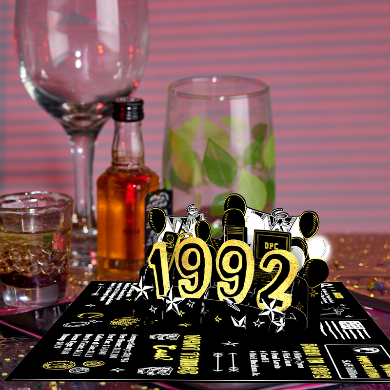 A 3D pop-up card featuring 1992 in gold glittery letters with retro-themed elements, surrounded by glasses, a small bottle of liquor, and party decorations on a table with a striped pink background