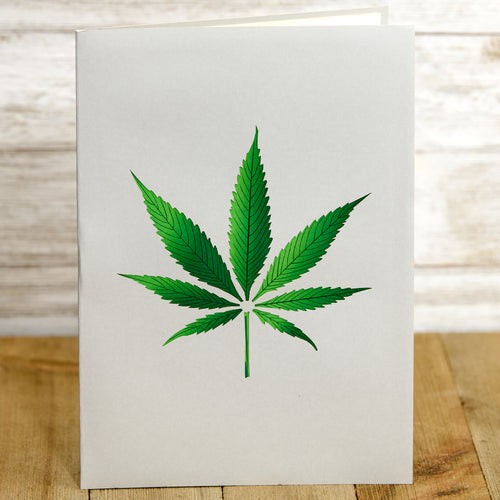 A greeting card with a green cannabis leaf illustration on a plain white background. The card is standing upright on a wooden surface, highlighting the simplicity and bold design of the leaf.