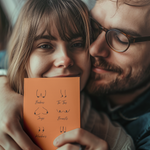 A smiling couple embraces while the woman holds a brown greeting card featuring simple line drawings of various breast shapes labeled with humorous terms like Boobies, Ta-Tas, and Jugs.